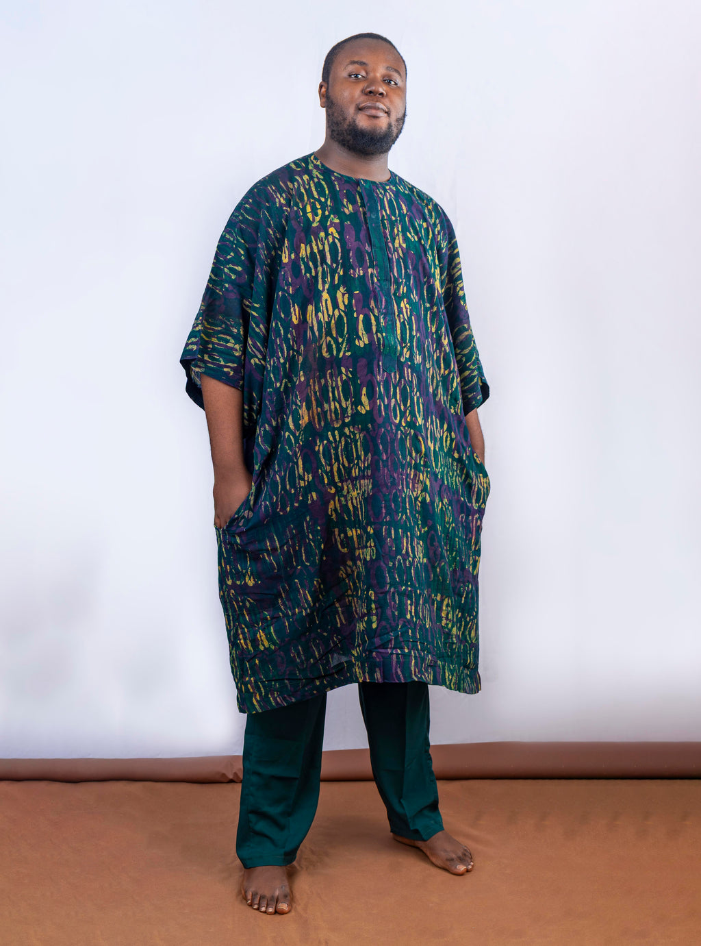 A man wearing a green, purple, and yellow abstract patterned African tunic shirt and dark green pants, standing against a white background.
