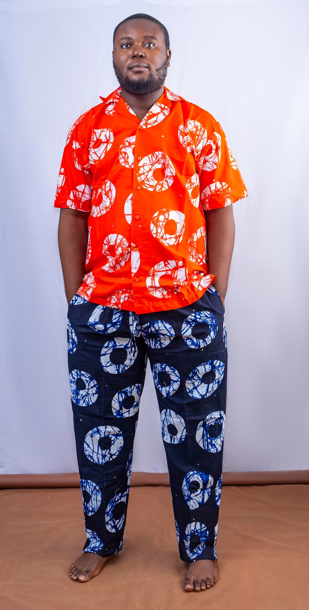 A man wearing a short-sleeved orange shirt with white circular patterns and dark blue pants with blue and white circular patterns.