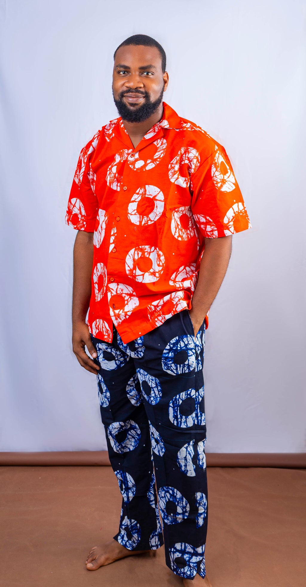 A man wearing a vibrant red short-sleeved shirt with white circular patterns and dark blue pants with white and blue circular patterns, posing casually with one hand in his pocket.