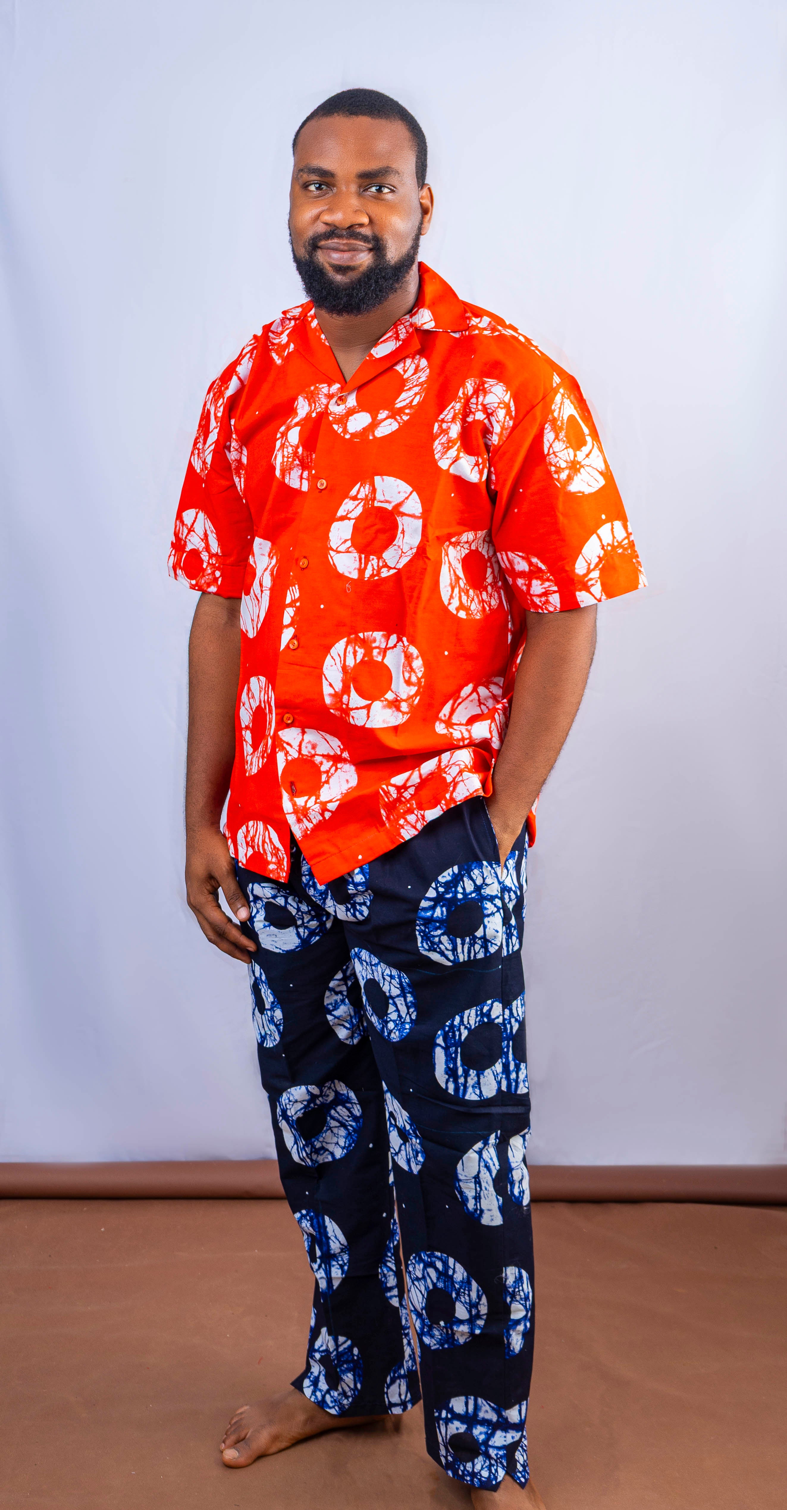 A man wearing a vibrant red short-sleeved shirt with white circular patterns and dark blue pants with white and blue circular patterns, posing casually with one hand in his pocket.