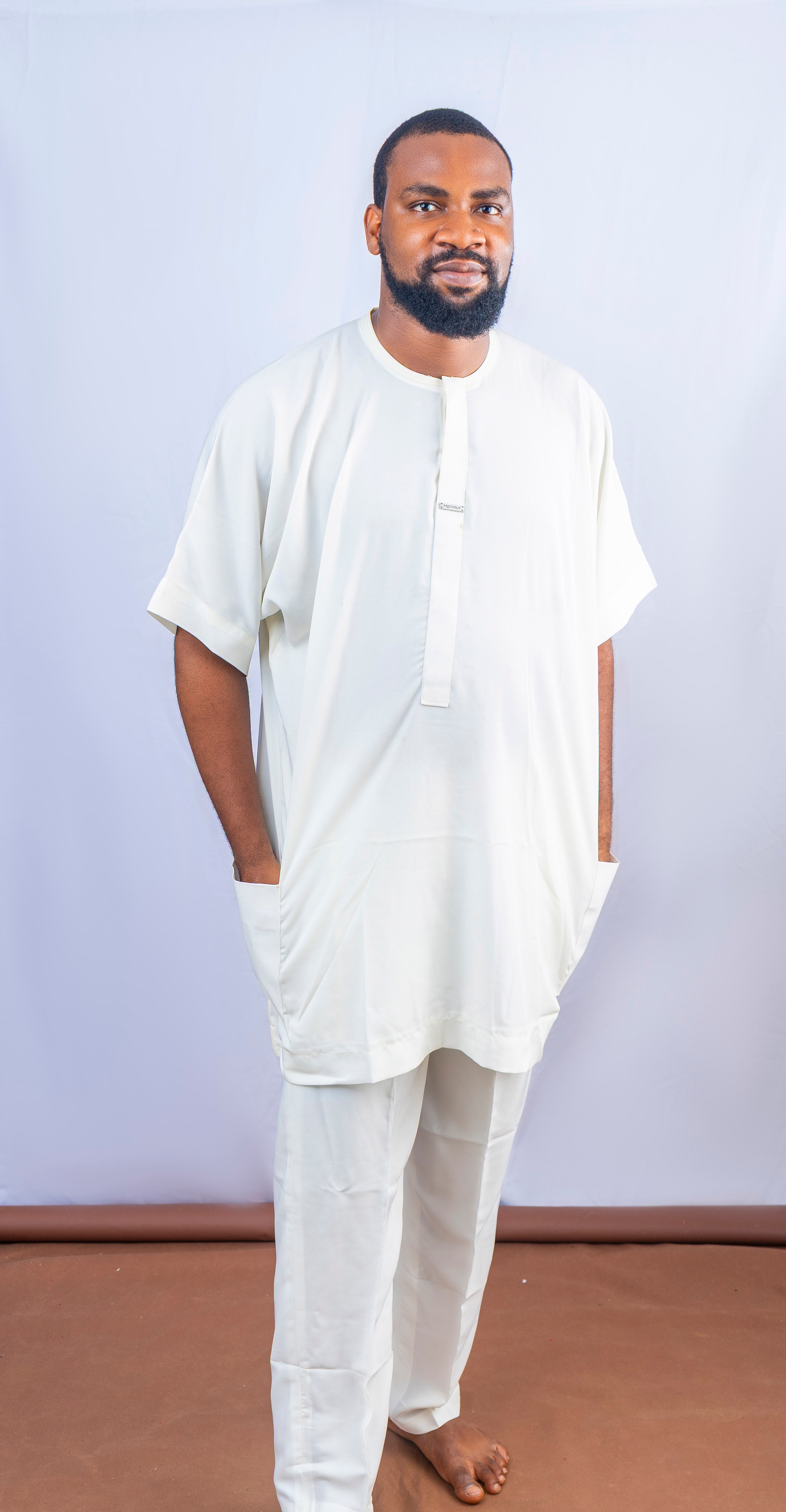 A man wearing a white traditional kaftan top with short sleeves and matching pants, posing confidently with hands in pockets.