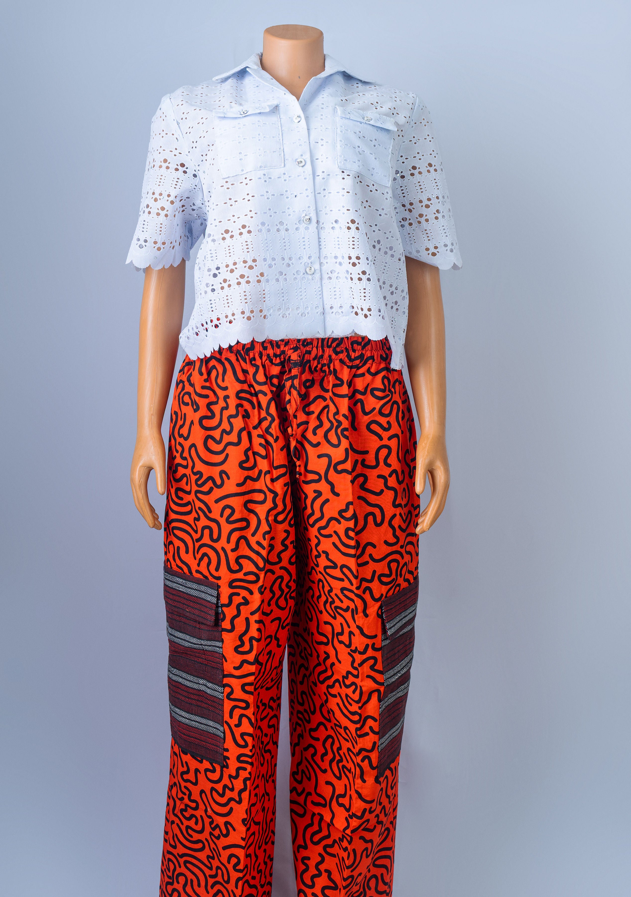 Mannequin wearing a short-sleeved white eyelet collared shirt with a scalloped hem, tucked into vibrant orange cargo pants with an abstract black squiggly pattern and striped utility pockets.