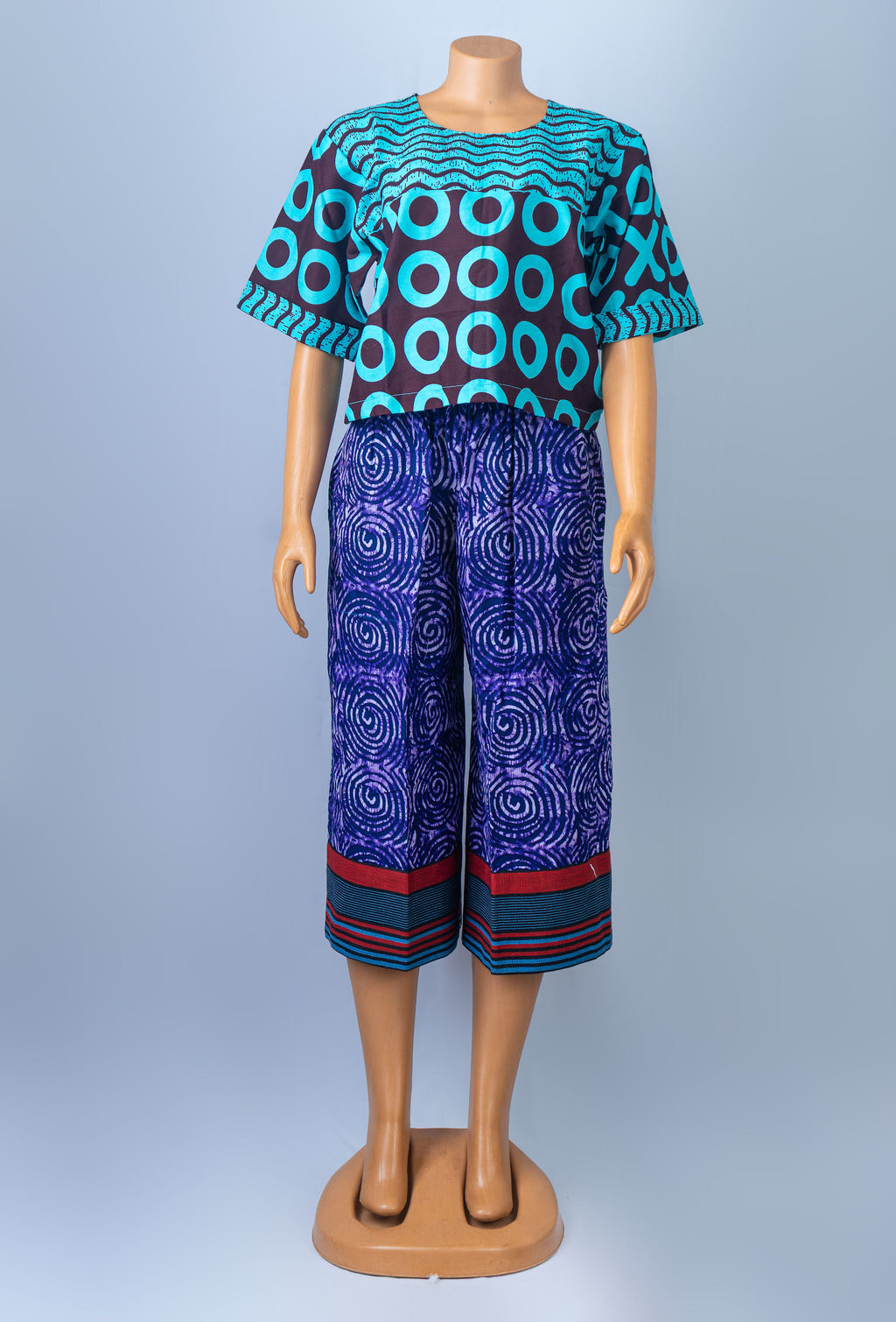 Mannequin modeling a colorful African print two-piece outfit, consisting of a short-sleeved top with teal and brown geometric circles and purple capri pants with a swirling pattern and striped cuffs.