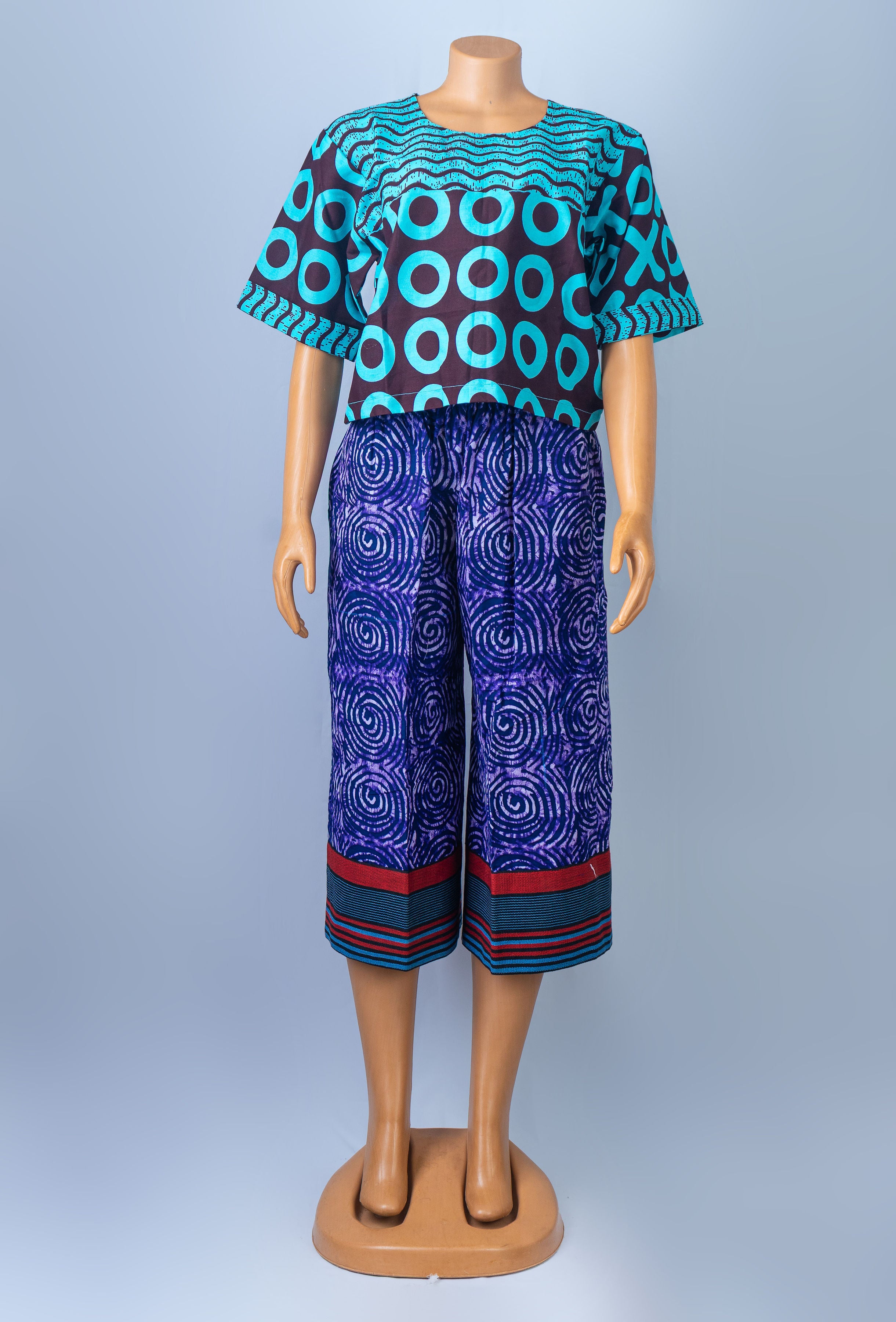 Mannequin modeling a colorful African print two-piece outfit, consisting of a short-sleeved top with teal and brown geometric circles and purple capri pants with a swirling pattern and striped cuffs.