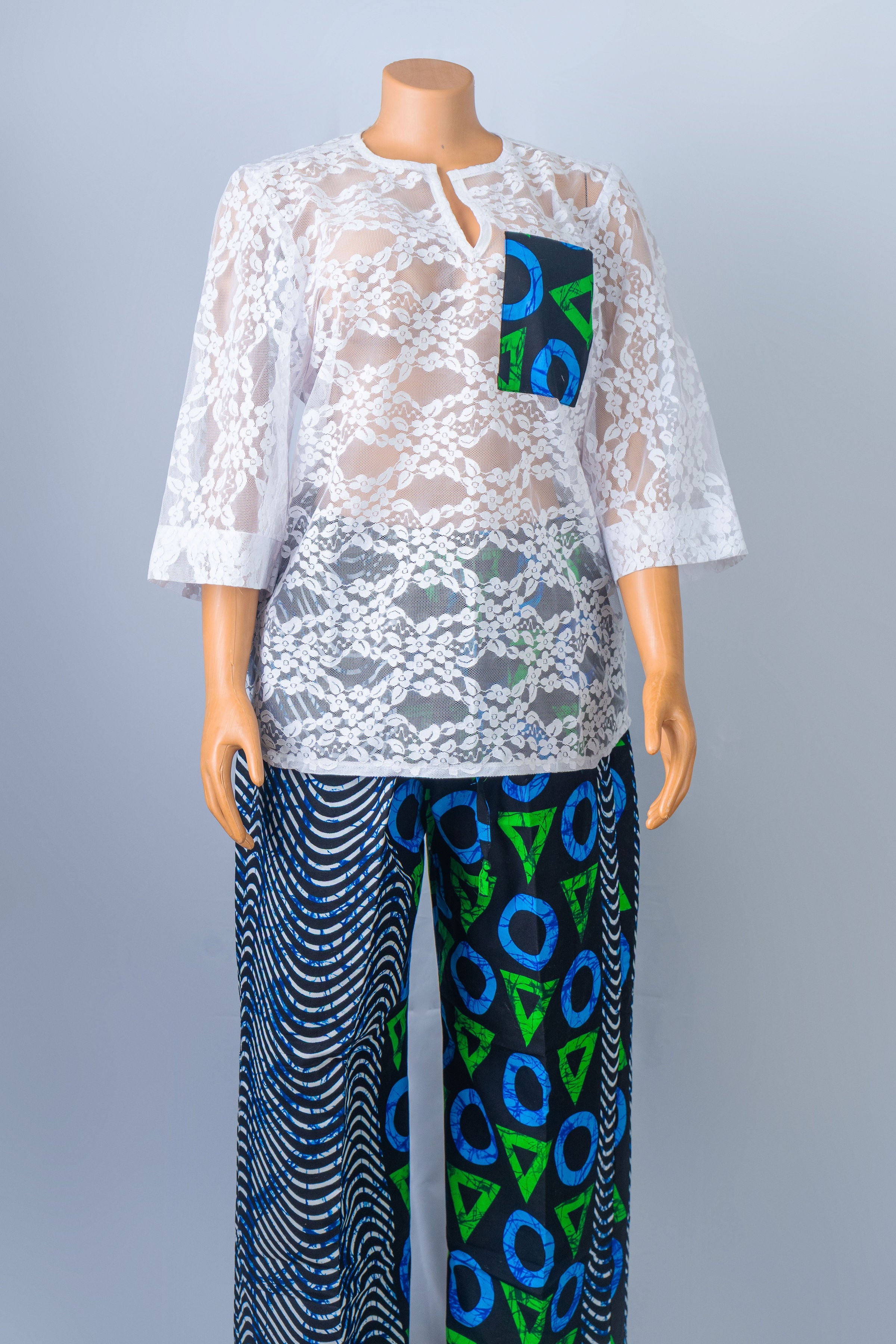 Mannequin wearing a white lace long-sleeve top with a colorful patterned pocket, paired with blue and green geometric patterned wide-leg pants.